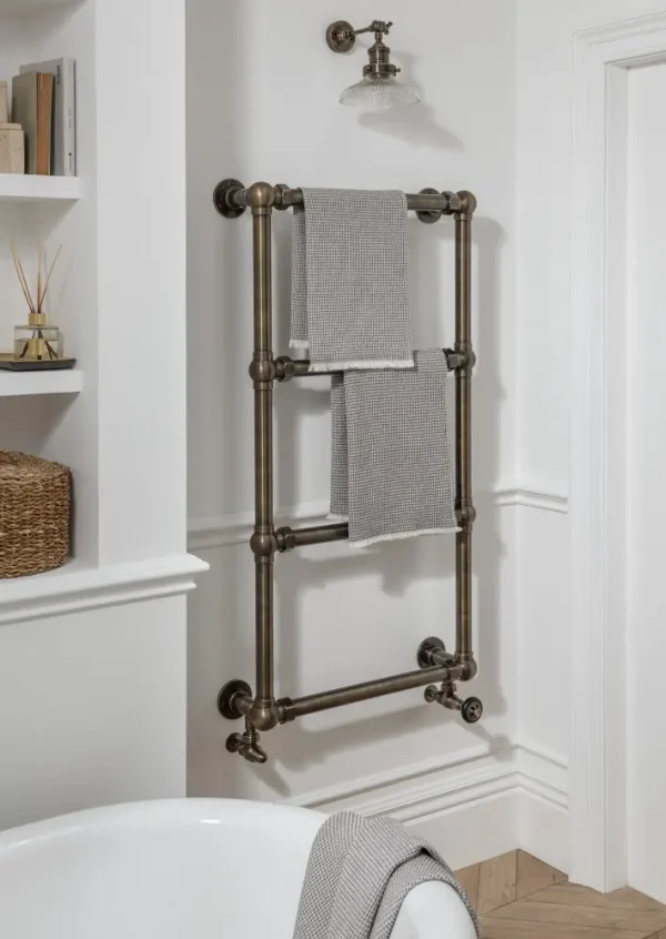 The 4 Bar Audley Heated Towel Rail, floor and wall-mounted, features two gray towels beside a white bathtub and decorative shelves.