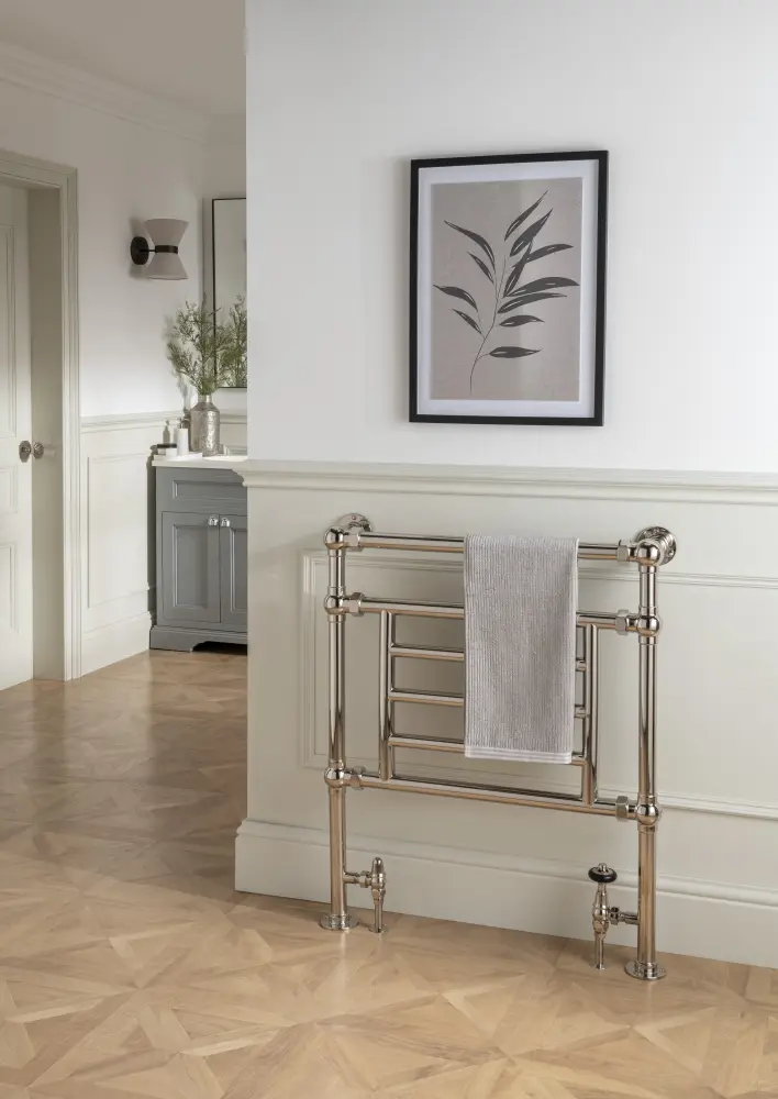 A bathroom scene showcases the 4 Bar Audley Heated Towel Rail with a towel, framed leafy artwork on the wall, and part of a vanity with a plant, all atop a light wood floor.