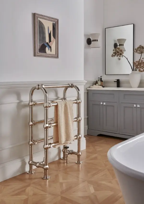 A bathroom features the Manton 7 Bar Heated Horse Towel Rail with a beige towel, gray vanity with mirror, wall sconce, framed abstract art, and light wood parquet flooring.