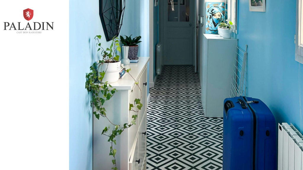 A narrow corridor with blue walls, geometric black-and-white floor tiles, potted plants on a white cupboard, a blue suitcase, and artwork near the door.