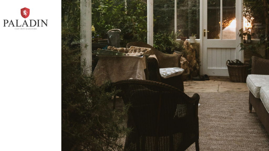 A cosy indoor patio with wicker chairs, a sofa, a table, potted plants, and a glass door leading outside, shown beside a Paladin Cast Iron Radiators logo.