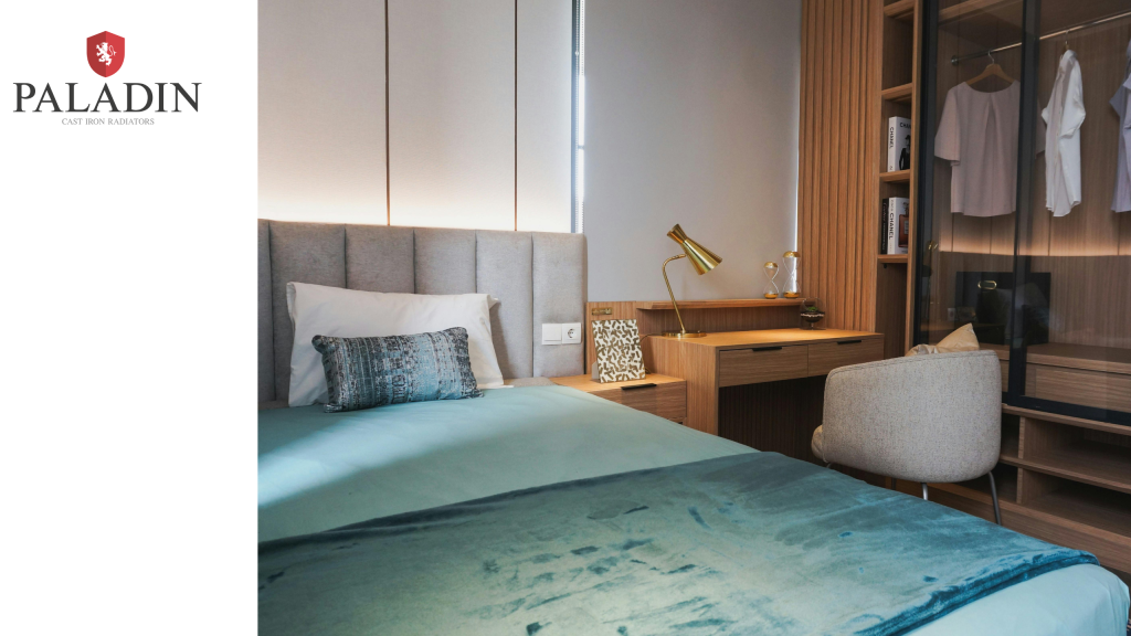Modern bedroom with a neatly made bed, a pillow and teal blanket, a desk with a lamp and chair, a wardrobe with hanging clothes, and natural lighting. Paladin logo in top left corner.