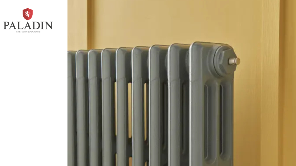 A grey cast iron radiator is mounted against a yellow wall. The Paladin logo and text appear in the top left corner on a white background.