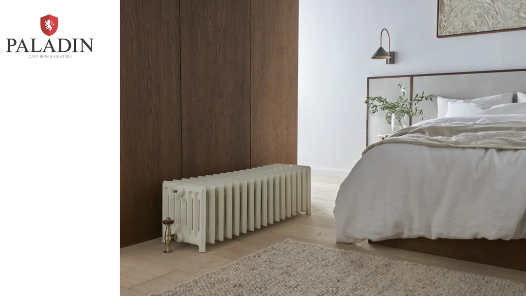 A white cast iron radiator sits beside a wooden partition in a modern, minimal bedroom with a neatly made bed and neutral décor. Paladin logo appears on the left.