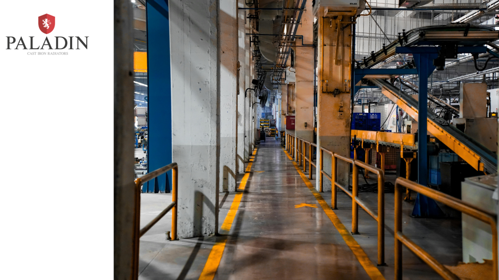 Industrial factory interior with metal railings and machinery along a central walkway; Paladin Cast Iron Radiators logo displayed on the left-hand side of the image.